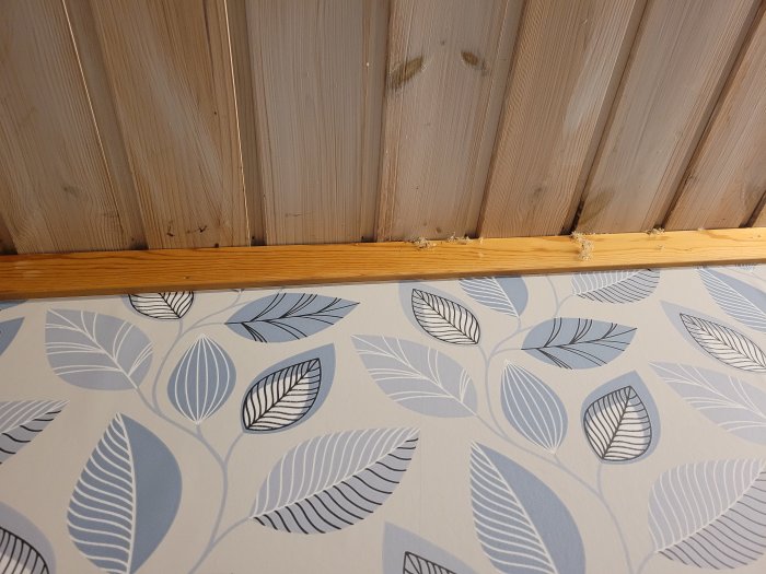 Träpaneltak möter blåvitt blad-mönstrad tapet med synlig träregel och fogskum.