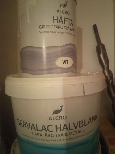 Två burkar med Alcro-färg, en med grundfärg och en med halvblank lackfärg, bredvid en borrspiral.