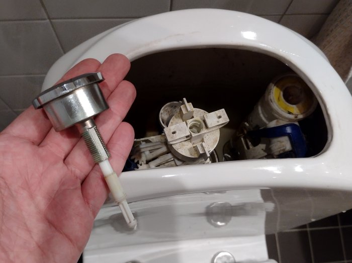 Händer som håller en toalettspolningsmekanism ovanför en öppen vattentank på en toalett.