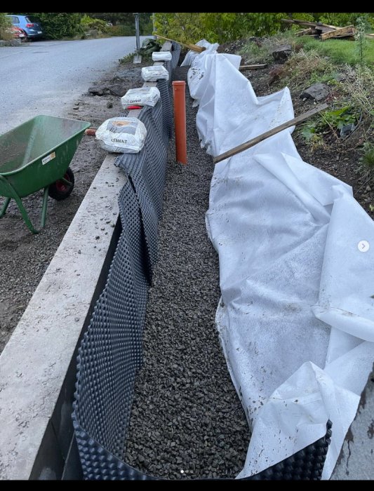 Avloppsrör och dräneringsarbete vid en mur med markduk, grus och säckar med cement.