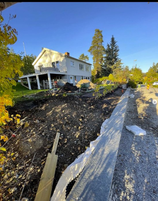 Ett pågående trädgårdsarbete med synlig dränering och nyplanterade syrener vid ett hus med en veranda.