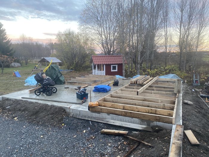 Grund och påbörjad trästomme för garage med gräsmatta och lekstuga i bakgrunden vid skymning.