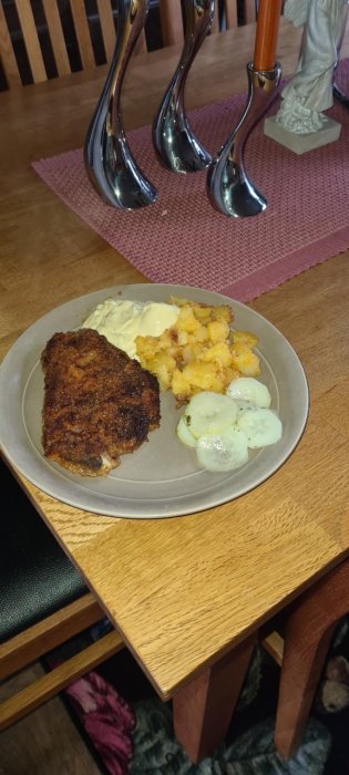Tallrik med panerad fläskkarré, stekt potatis med lök, skivad gurka och bearnaisesås på ett matbord.