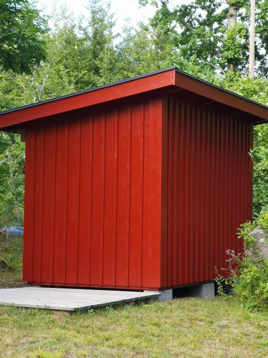 Röd trädgårdsbod med lockläktpanel och platt tak mot grönskande bakgrund.