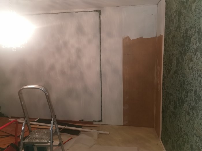 Renoveringsarbete i ett rum med exponerade spånskivor, vita väggar och gammal tapet, med en stege och byggmaterial på golvet.