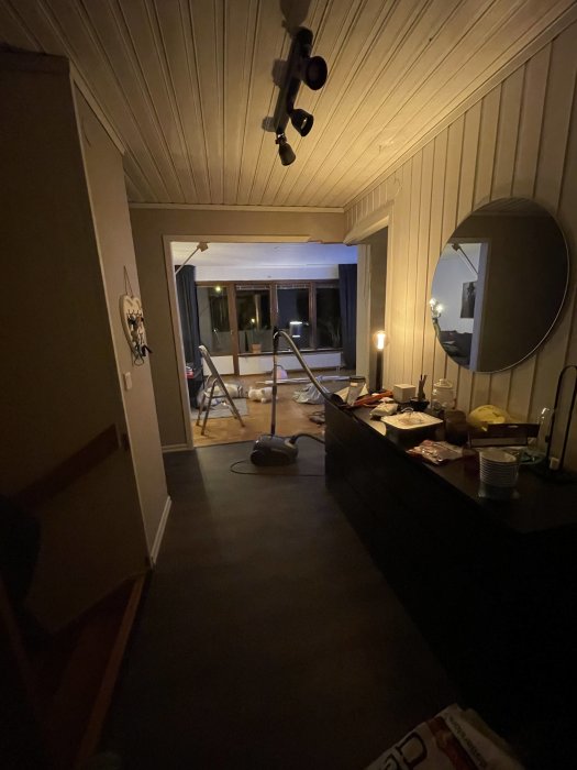 Ett rum under renovering med öppen vägg mot vardagsrum, byggmaterial och verktyg synliga.