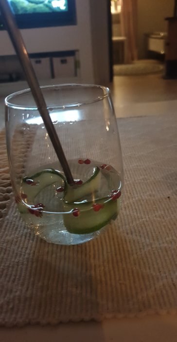 Glas med Gin och Tonic med skivad gurka och rosépepparkorn, bakgrund av suddigt vardagsrum.