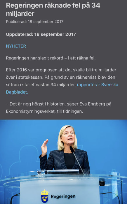 Kvinna talar vid podiet med texten "Regeringen" och en mikrofon, med blå bakgrund och logga.