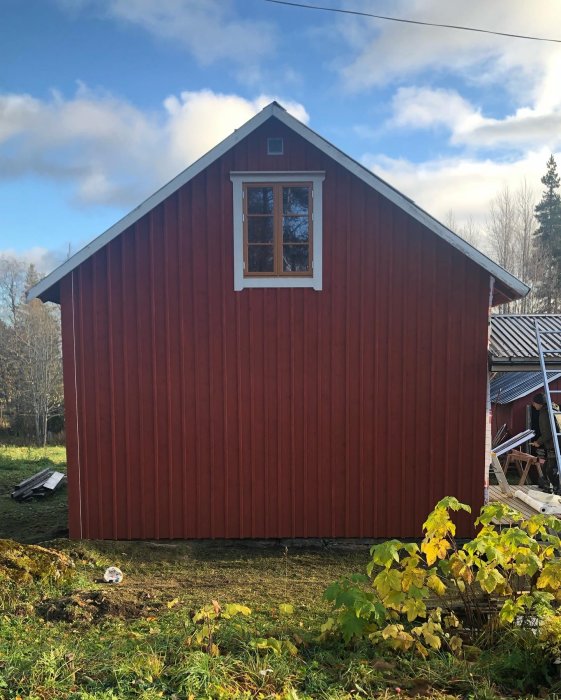 Liten fönster på en större röd trägavel av ett hus med grön växtlighet i förgrunden.