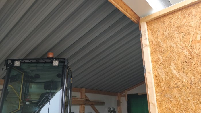 Inre hörn av ett garage med grå plåttak och OSB-skivor på väggen, del av en grävmaskin syns.