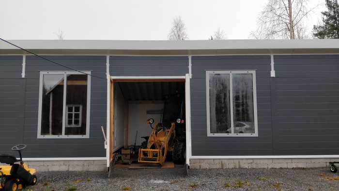 Mörkgrått garage med öppen port som visar en gul hjullastare, flankerat av två fönster mot en grusgård.