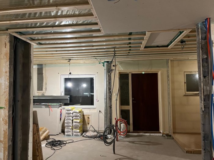 Renoveringsprojekt med osluttede väggar, gipsplattor och synliga rör i taket, samt byggmaterial på golvet.