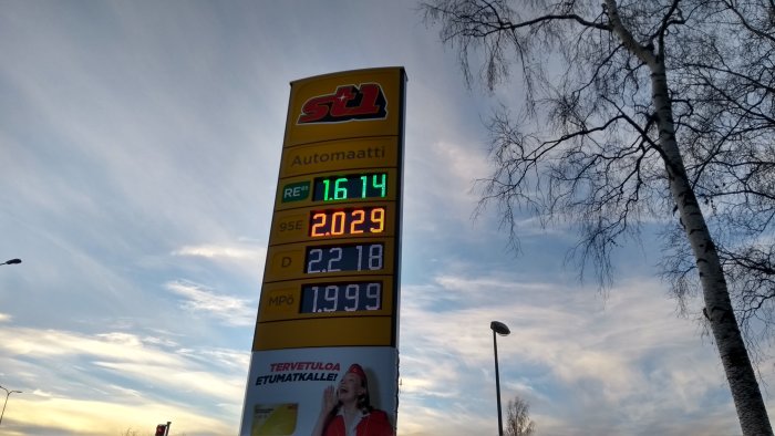 Bränslepristavla i Finland med priser för bensin och diesel, reklamskylt och björkträd mot himmel.