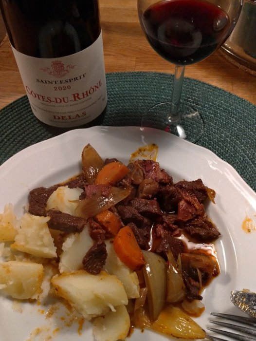 En tallrik med köttgryta, morötter och potatis, bredvid ett glas rött vin och en vinflaska.