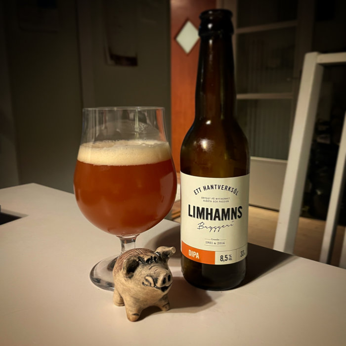 Ett glas öl och en flaska med etiketten "Limhamns Bryggeri", framför en liten gristräfigur på ett bord.