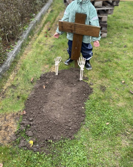 Barn håller i ett träkors framför en jordhög med två dekorativa skeletthänder som sticker upp ur marken.