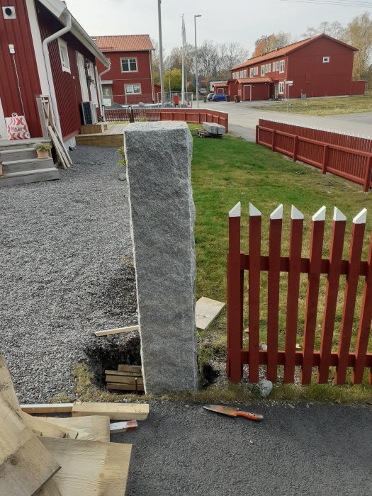 Stor stenstolpe placerad intill ett rött trästaket med arbetsmaterial runtomkring på en grusad tomt.