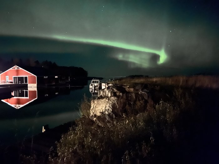 Nordljus över natthimmel, med reflektion i vatten vid stuga och båt.