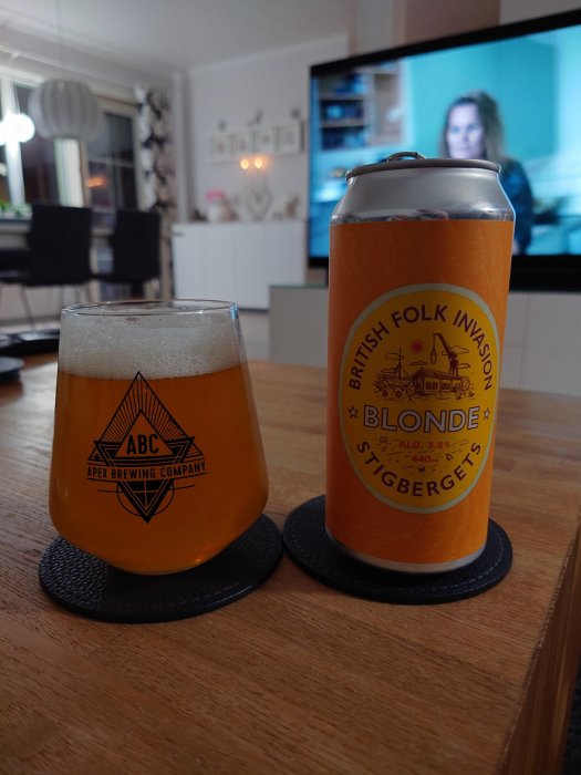 Ölglas och burk från Apex Brewing på ett bord med en oskärpt TV i bakgrunden.