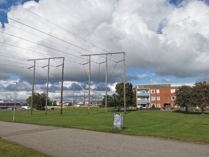 Bostadsområde med högspänningsledningar nära flerfamiljshus och grönområde under molnig himmel.