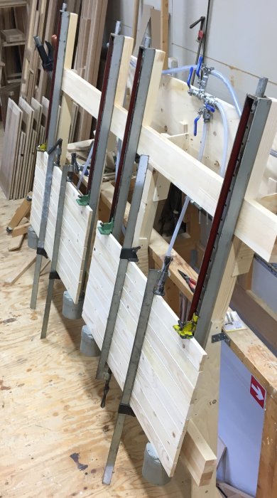 Trähantverk i en verkstad: limmade träpaneler fastklämda för torkning mot bakgrund av verkstadsverktyg och timmer.