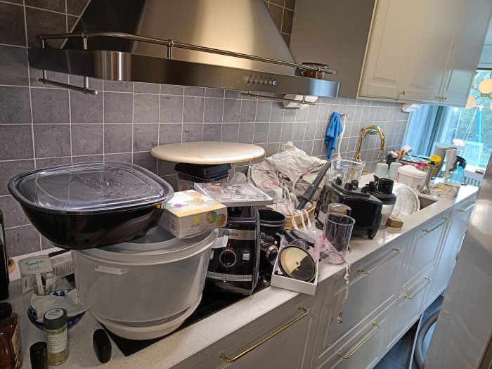 Köksbänk full av renoveringsmaterial och köksutrustning under en fläkt och grå kakelvägg.