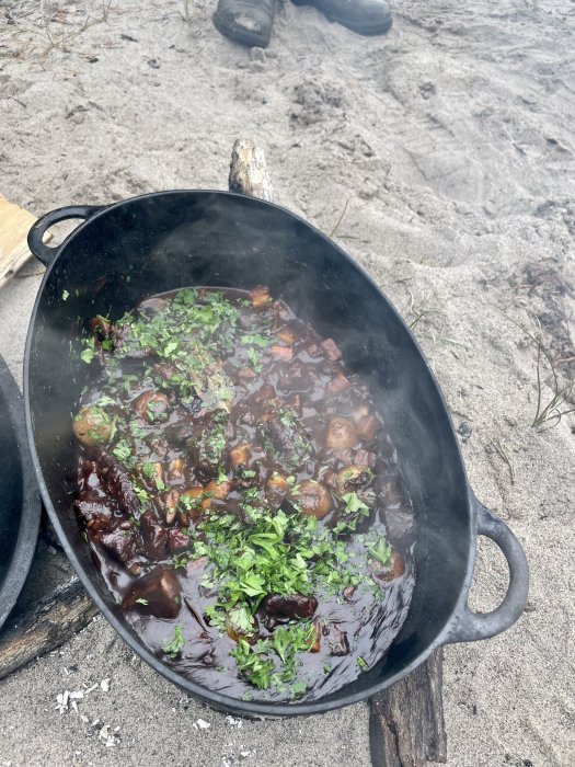 Järngryta med boeuf bourguignon tillagad på strand över öppen eld, garnerad med örter.