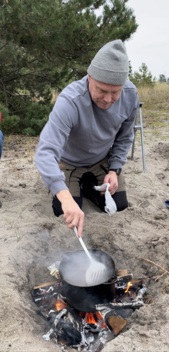 Person lagar mat i järngryta över öppen eld på en strand.