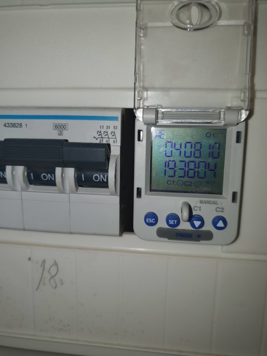 Digital timer för ytterbelysning fastsatt på vit vägg, visar klockslag och programmeringsknappar.