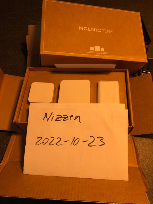 Öppen kartong med texten "NGENIC TUE" och flera enheter, framför ett handskrivet papper med datum "2022-10-23".