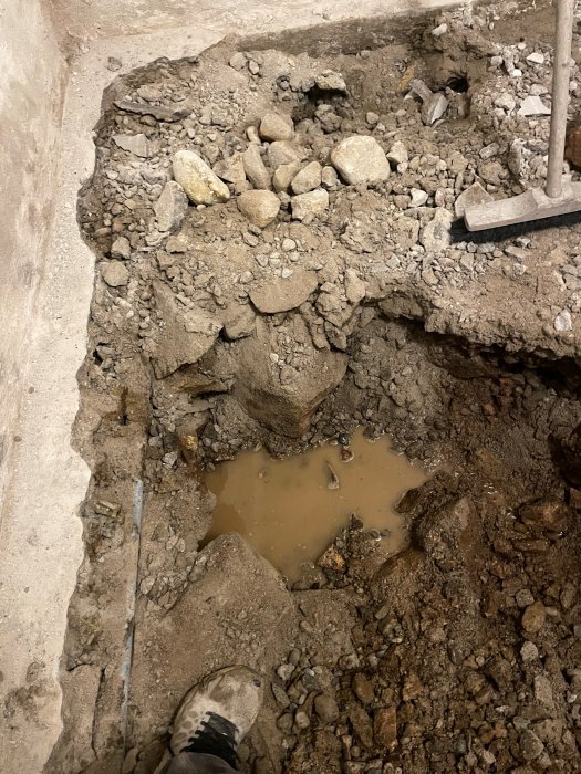 Uppbruten betonggolv med grus, stenar och en vattenpöl, spade och en persons skotopp synlig.