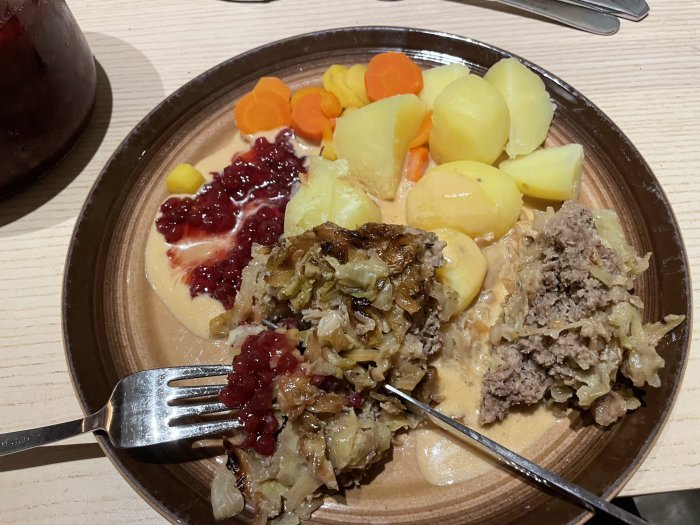 Hemlagad kålpudding serverad med potatis, morötter, gräddsås och lingonsylt på en tallrik.