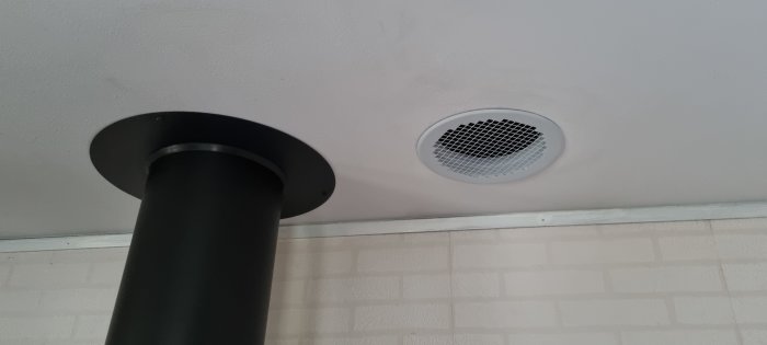 Ett svart isolerat flexrör i taket bredvid en vit ventilgaller på en vitmålad vägg.