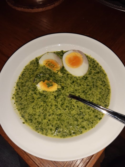 Skål med grön spenatsoppa och halverade kokta ägg, med sked, ovanpå träbord.