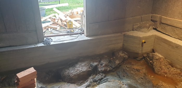 Grävd grop i ett uthus med berggrund, synliga dräneringskanaler och nygjutna betongväggar, arbetsmaterial i bakgrunden.
