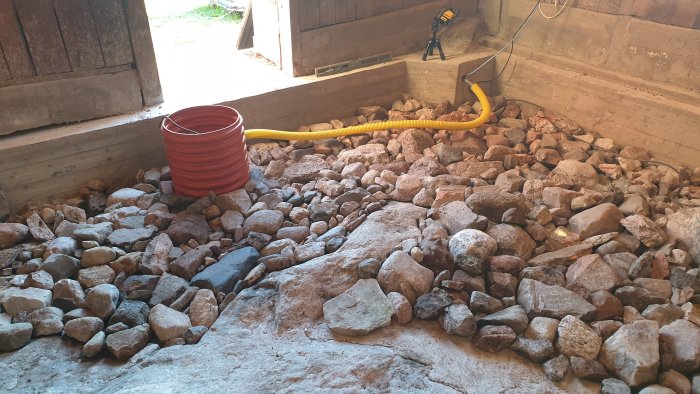 Grund av ett uthus fylld med sten och singel, synlig pumpgrop och dränageslang.
