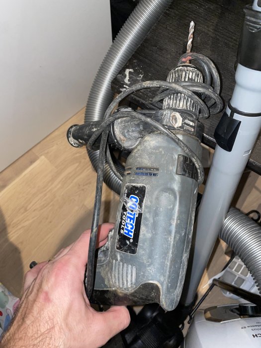 Hand håller en dammig och använd Cotech-vattenpump under ett köksskåp med synliga rör och slangar.