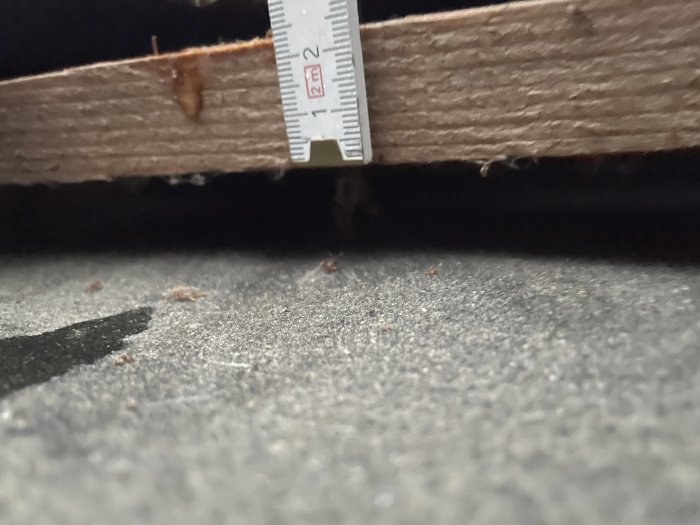 Mätning av avståndet under en träbjälke med tumstock som visar cirka 2 centimeter tomrum.