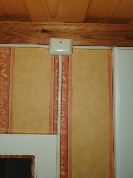 En elkabel dragen i kabelkanal längs med en taklist och in i en dosa ovanför en tapetserad vägg.