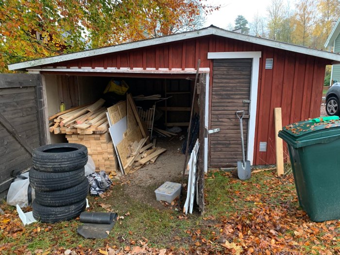 Öppet förråd med trävirke, bildäck och verktyg, omringat av löv och en soptunna utomhus under hösten.