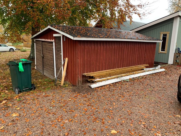 Röd träförrådsbyggnad med vita dörrar omgiven av fallna löv och med byggmaterial staplat vid sidan.