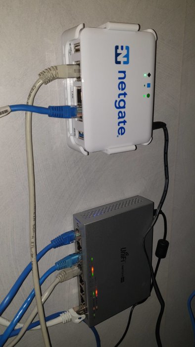 En Netgate 1100-router och UniFi US 8 60W switch med kopplade ethernetkablar på en vägg.