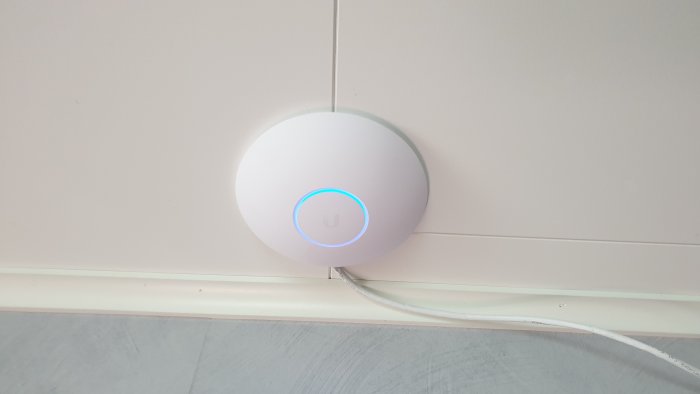 UniFi UAP-AC-LR accesspunkt monterad på vägg med synlig Ethernet-kabel och blått ljus.