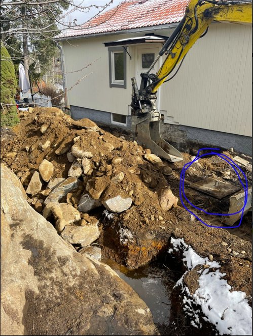 Grävmaskin vid gammal brunnsplats nära hus med synligt vatten och stenar, markarbete pågår.