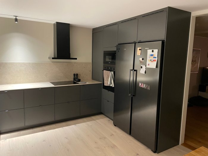 Modernt kök med grå skåp, inbyggda apparater och kyl med anslagstavla samt avvaktande belysning.