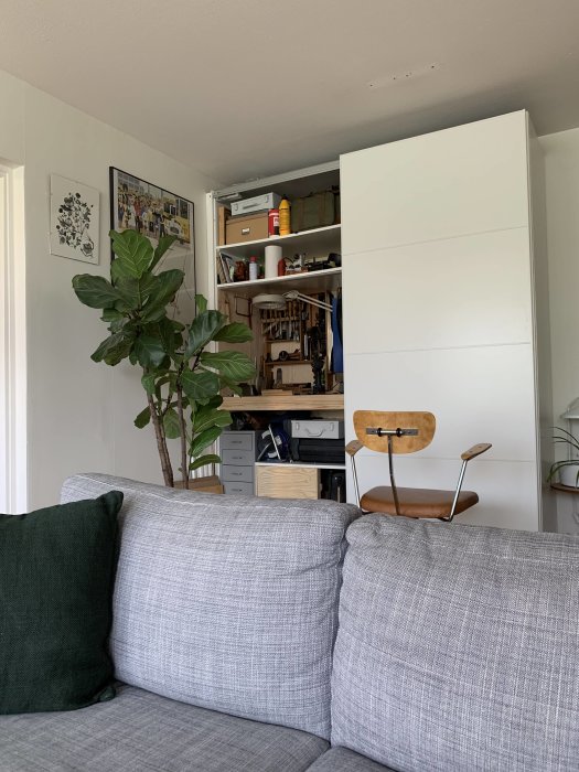 Vardagsrum med en öppen IKEA garderob som innehåller verktyg och material, bredvid en soffa och en stol.