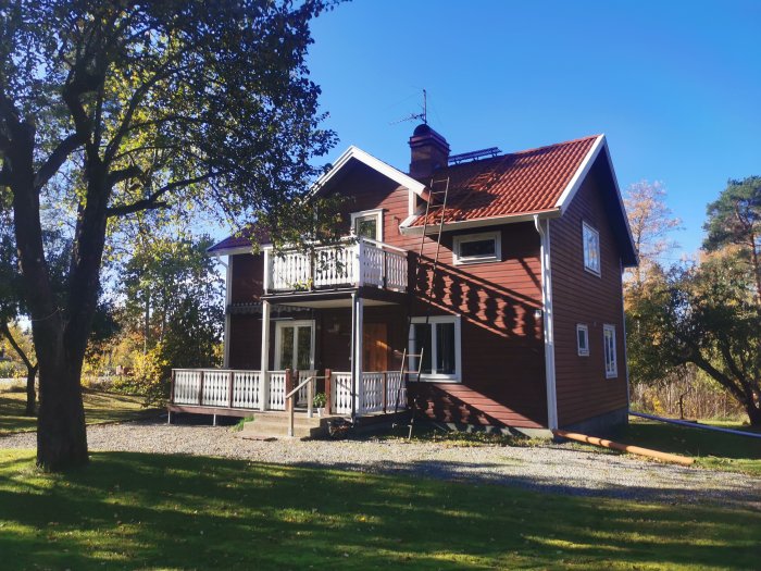 Tvåplans trähus med röd fasad och vita fönster, veranda, omgiven av grönskande trädgård.