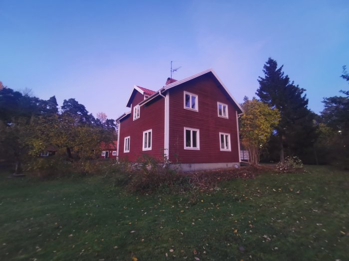 Röd tvåvåningshus i skymning med grönskande trädgård och startklar anten på taket.