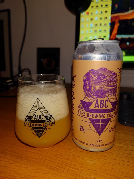 Glas med öl och en ölburk från Apex Brewing Company på ett skrivbord, bakgrund med datormonitor.