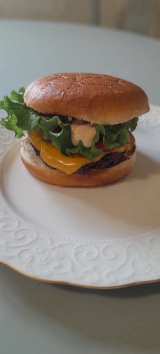 Hemlagad hamburgare med sallad, cheddarost, tomat och dressing på ett vitt mönstrat fat.